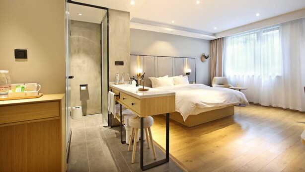 Une chambre d’hôtel moderne avec un plancher en bois, un grand lit avec du linge de maison blanc, un bureau avec une chaise, une douche en verre et une grande fenêtre avec des rideaux.