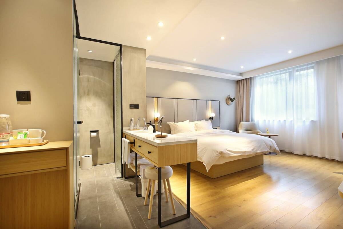 Een moderne hotelkamer met een houten vloer, een groot bed met witte lakens, een bureau met stoel, een douchecabine van glas en een groot raam met gordijnen.