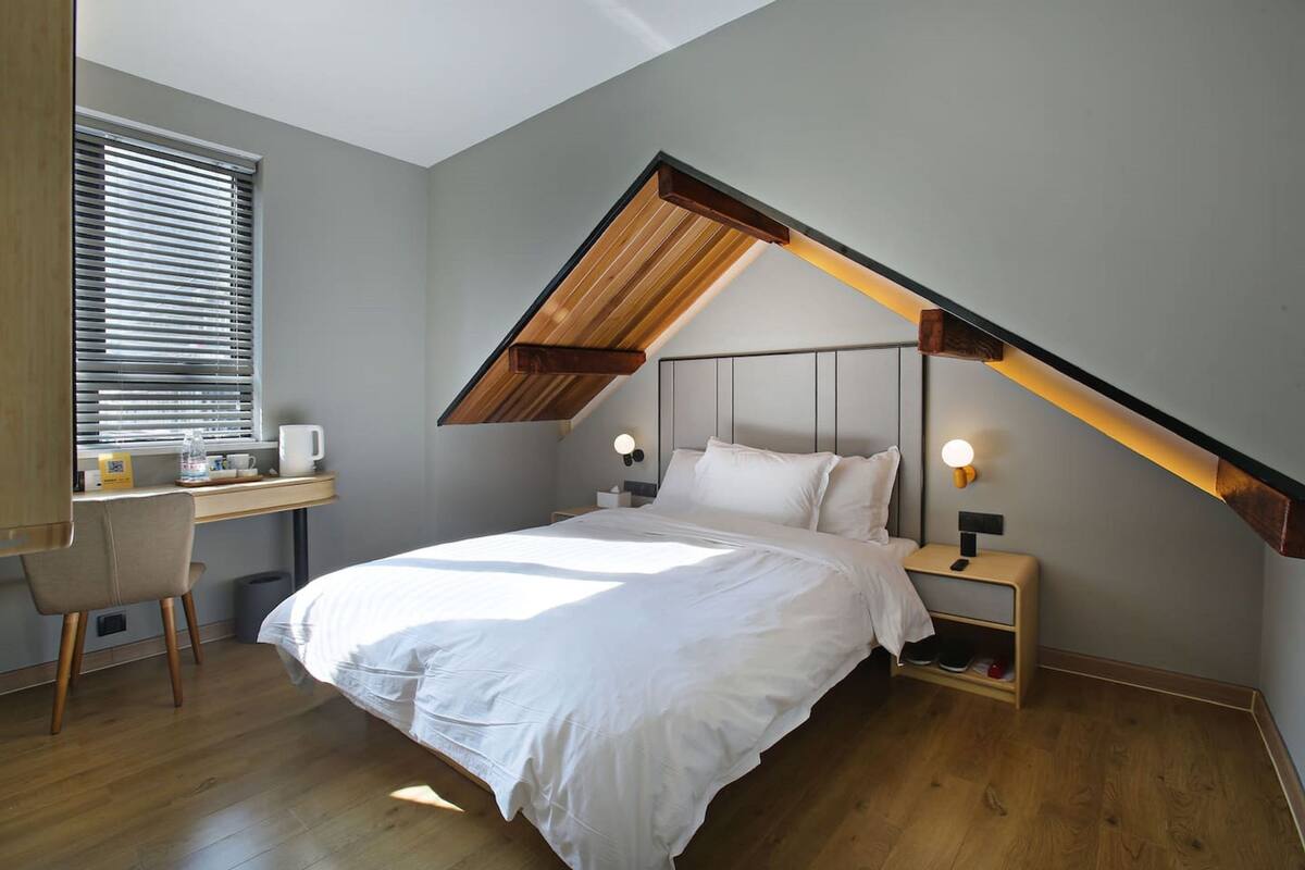 Een slaapkamer met een schuine plafond, een bed, nachtkastjes met lampen, een bureau en een stoel.