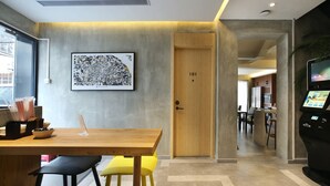 Une chambre d’hôtel moderne avec un bureau en bois, une chaise jaune, une œuvre d’art abstraite encadrée, une porte en bois portant le numéro 101 et un écran numérique sur le côté droit.