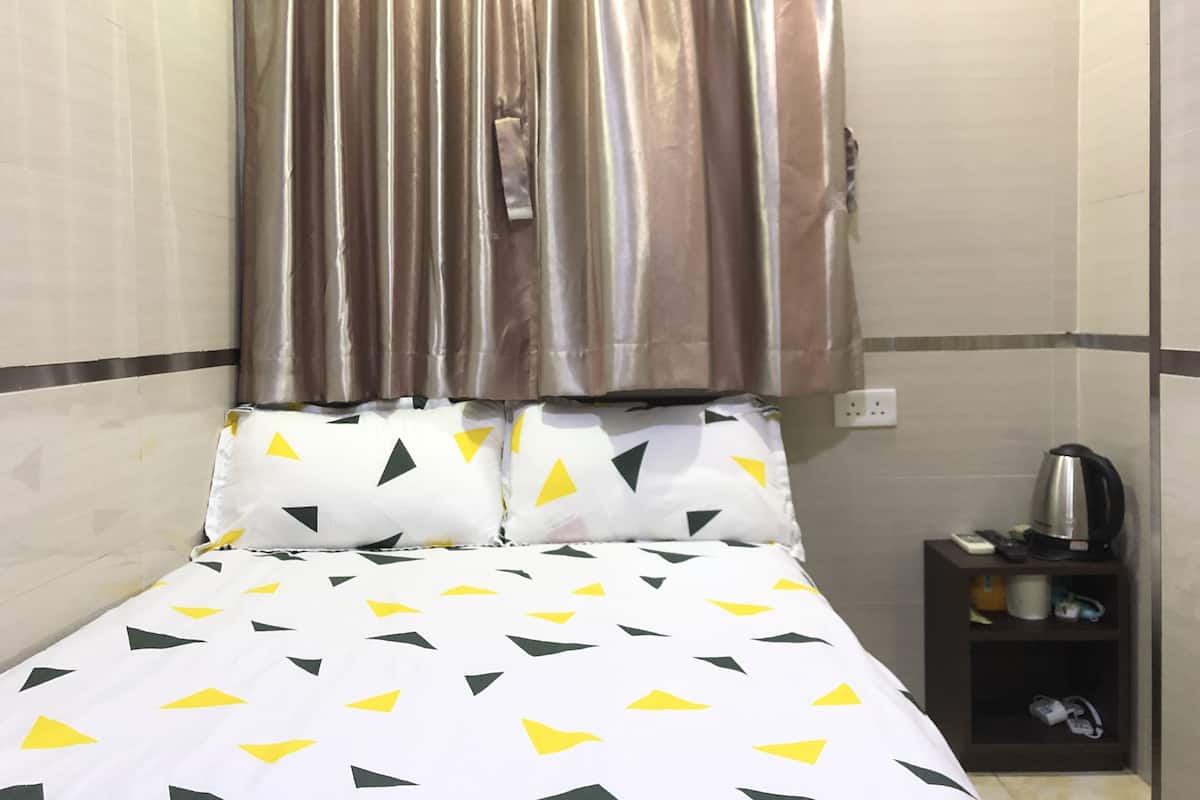 A hotel room with a bed, curtains, a nightstand with a kettle, and a wall socket.