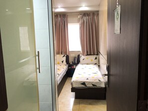 Quarto de hotel compacto com duas camas de solteiro, uma secretária pequena, chaleira e janela com cortinas.