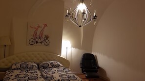 A bedroom with a bed, a chair, a chandelier, and a wall art of giraffes on a bicycle. - Proko 2 (Prague)