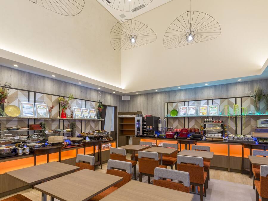 A modern restaurant with multiple dining tables and chairs, a buffet area with food displays, and decorative lighting fixtures.