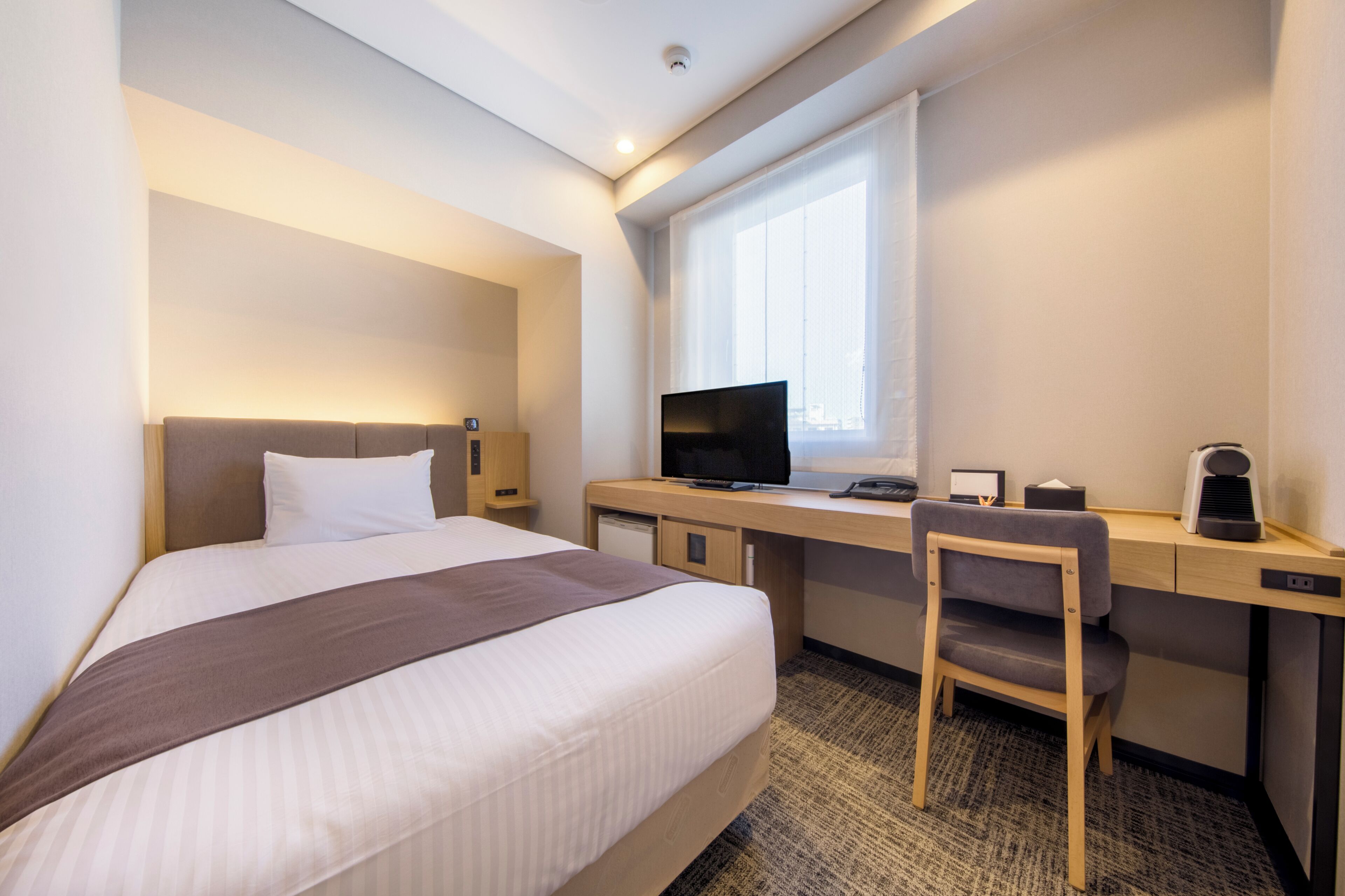 Ein Hotelzimmer mit einem Bett, einem Schreibtisch mit Computer, einem Stuhl und einem Fenster.