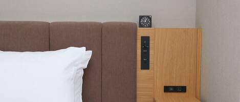 Una cama con cabecera, una mesita de noche con un reloj y un panel de control con botones etiquetados como