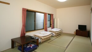 Een kamer met tatami vloer, twee tatami matten, een klein houten tafeltje, een televisie op een kast, en een raam met gordijnen.