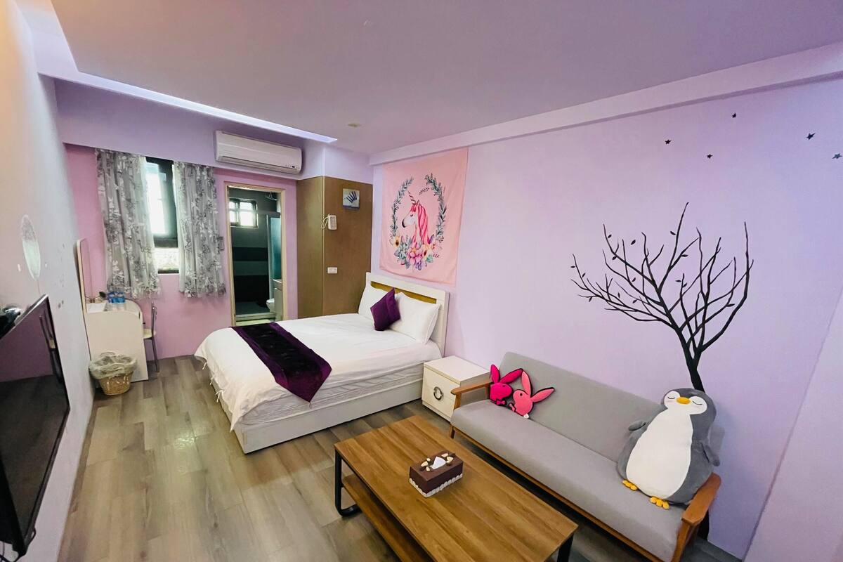 A hotel room with a bed, a sofa, a wooden table, a TV, and a bathroom visible through an open door.