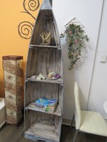 Una esquina con una estantería de madera que muestra libros y objetos decorativos, una silla blanca y una planta colgada en la pared.