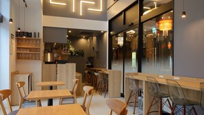 Um café moderno com mesas e cadeiras de madeira, área de cozinha visível e uma instalação luminosa decorativa na parede.