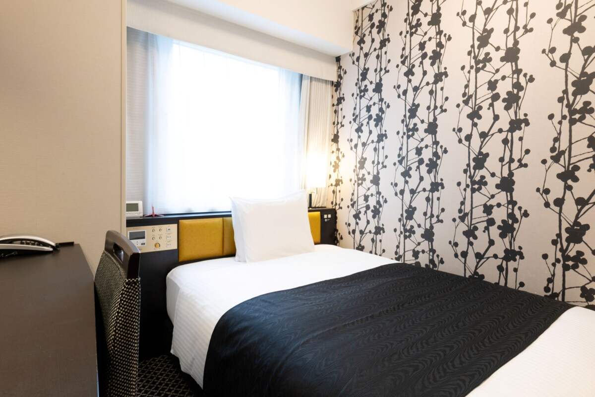 A hotel room with a bed, a bedside table, a lamp, and a window with curtains.