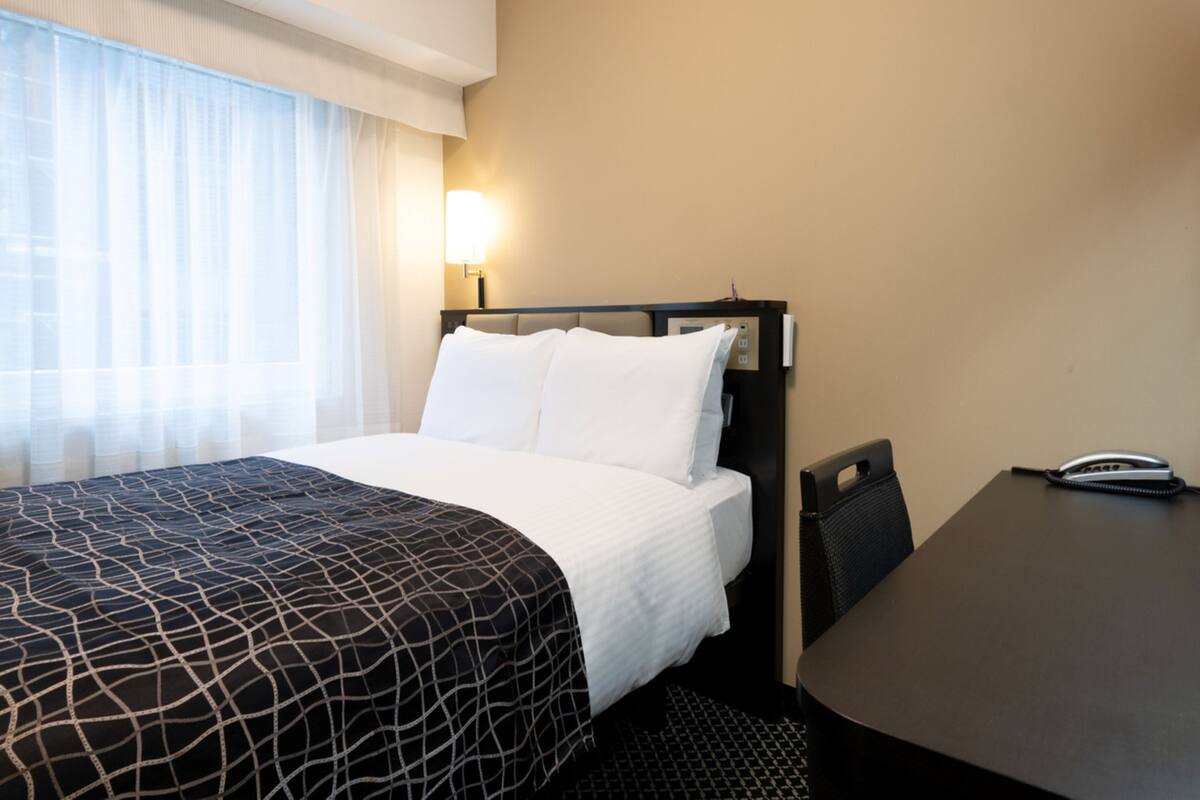 A hotel room with a bed, a desk, a chair, a lamp, and a telephone.