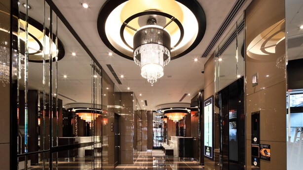 Um hall de hotel moderno, com pisos refletivos, um lustre central e paredes de vidro.