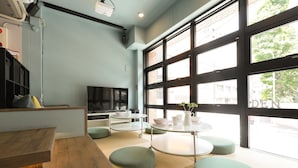 A modern interior with a wooden bench, a TV, and a seating area with round tables and green ottomans. - hostel DEN (Tokyo)