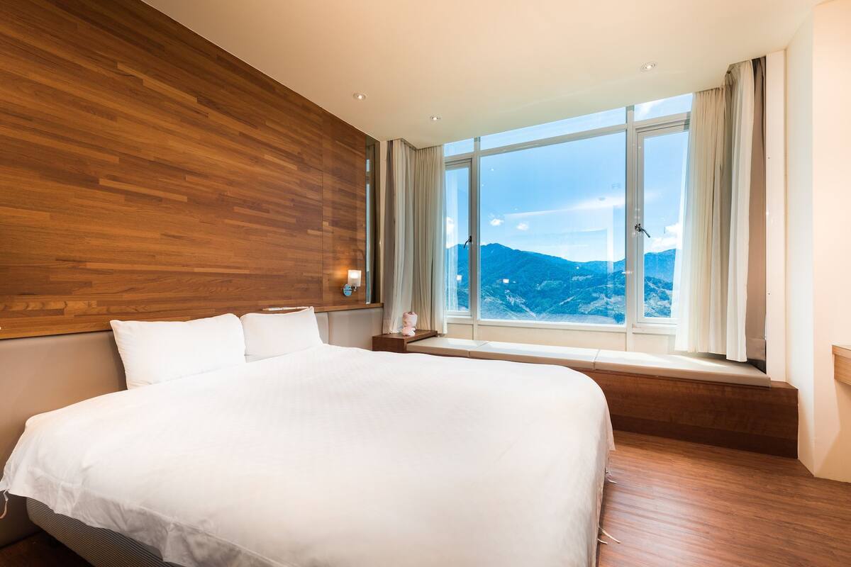 A hotel room with a large bed, wooden paneling, a window with a mountain view, and a bedside table with a lamp.