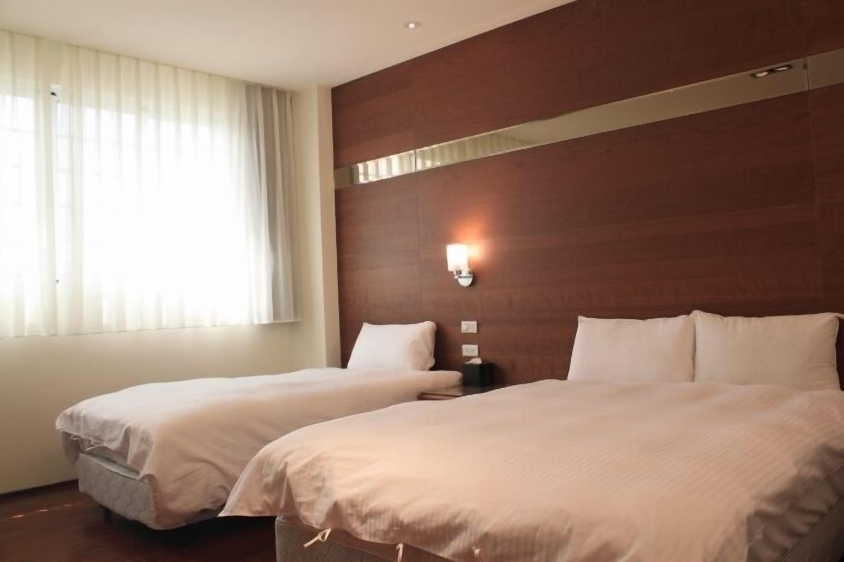 A hotel room with two beds, a wooden headboard, a wall-mounted light, and a window with blinds.