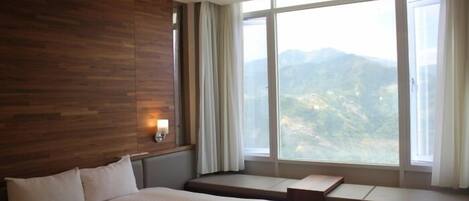 A hotel room with a large bed, a wooden accent wall, a window with a mountain view, and a bedside table with a lamp.