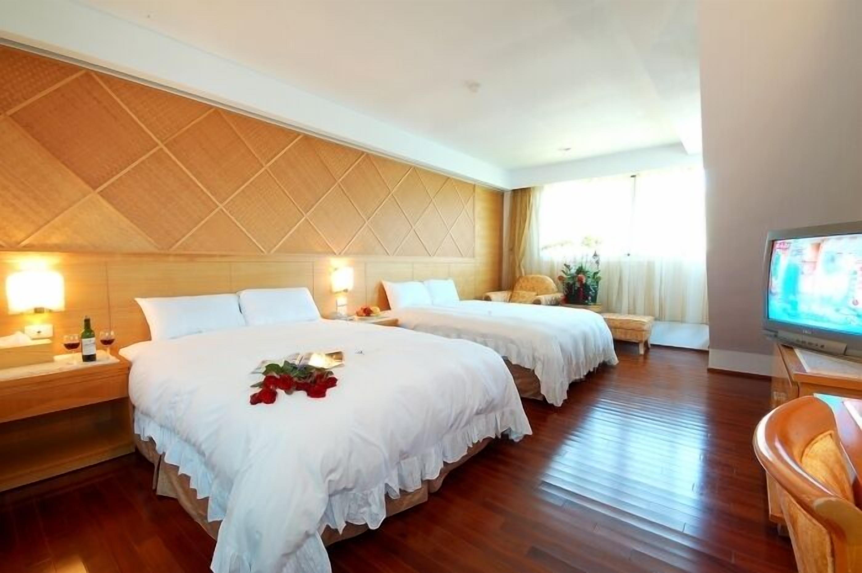 Habitación de hotel con dos camas, cabecero de madera, mesitas de noche con lámparas, un televisor y suelo de madera.