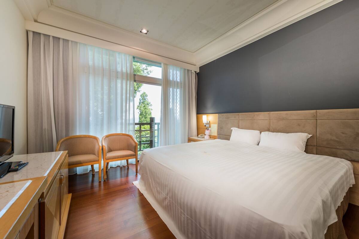 A hotel room with a large bed, a desk with a chair, a TV, and a window with curtains.