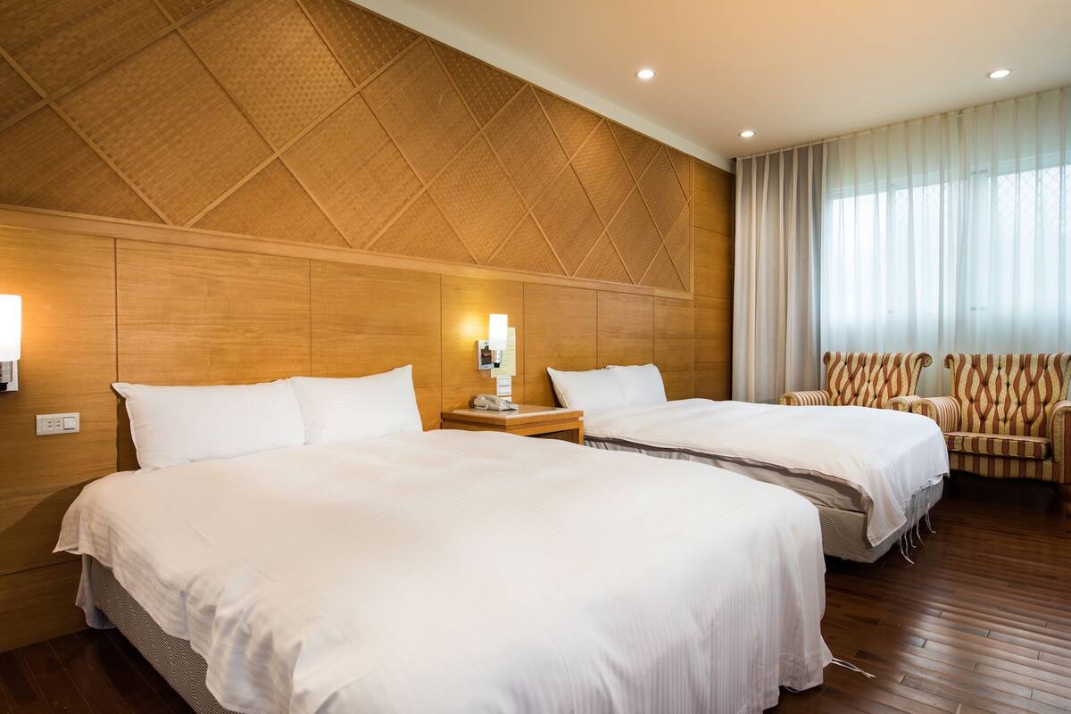 A hotel room with two beds, wooden paneling on the walls, a large window with curtains, and a chair with a striped pattern.