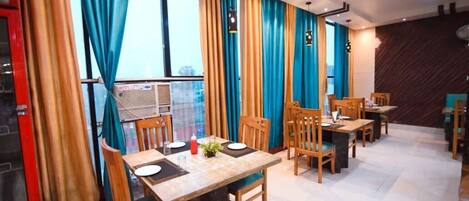 A dining area with wooden tables and chairs, large windows with blue curtains, and a modern ceiling design.