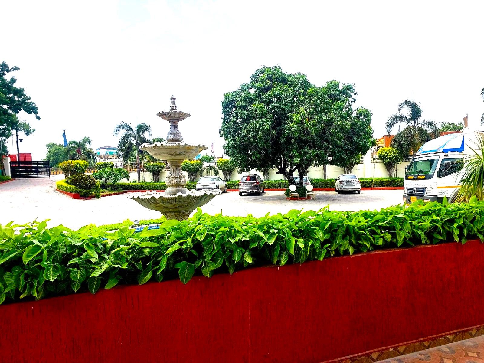 A fountain surrounded by greenery, a red wall, and parked cars.