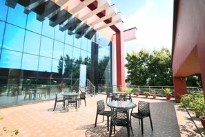 An outdoor seating area with tables and chairs, a glass building facade, and potted plants.