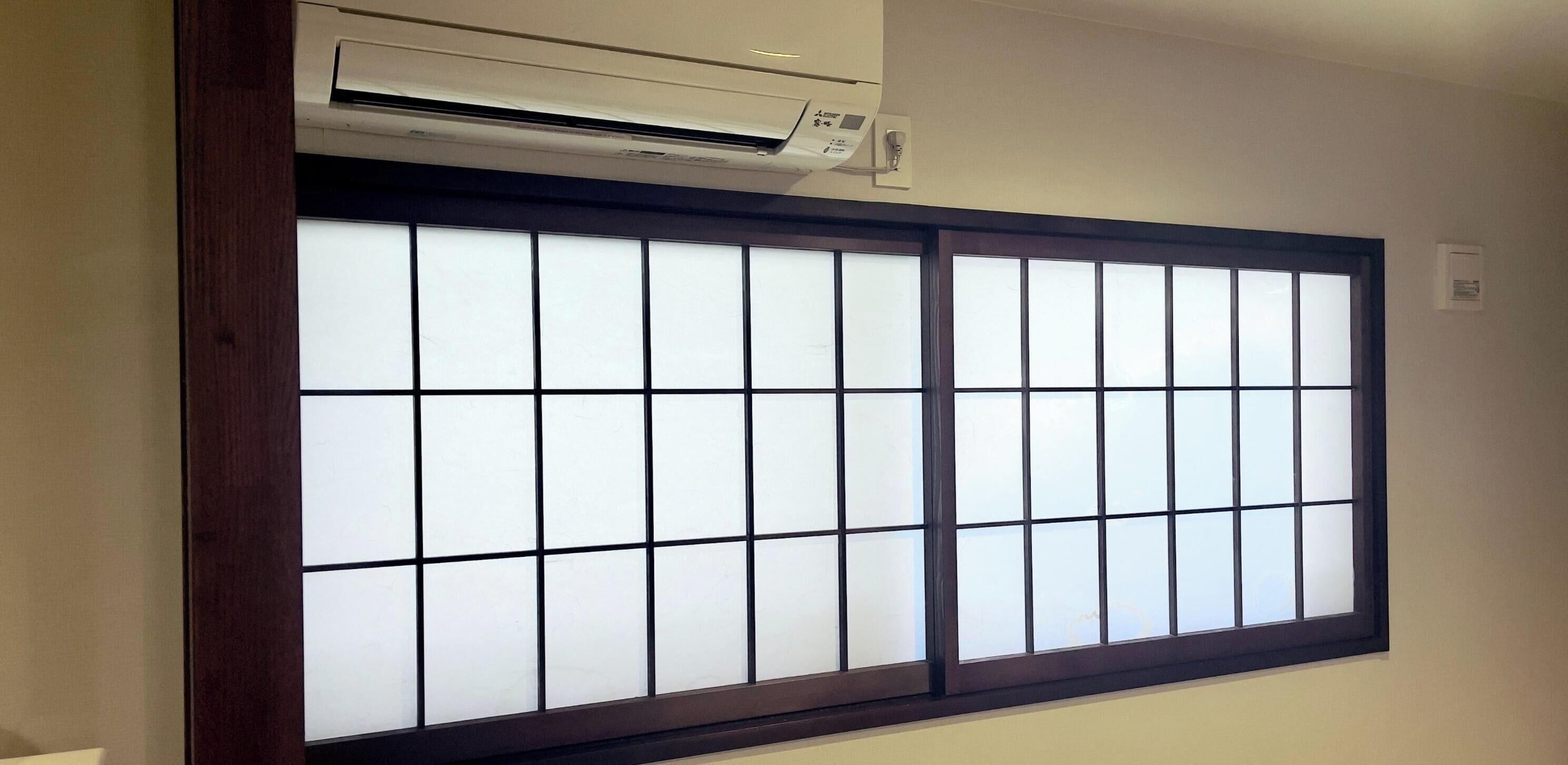 A room with a large window featuring a grid pattern, an air conditioning unit mounted above it, and a wall-mounted light switch.