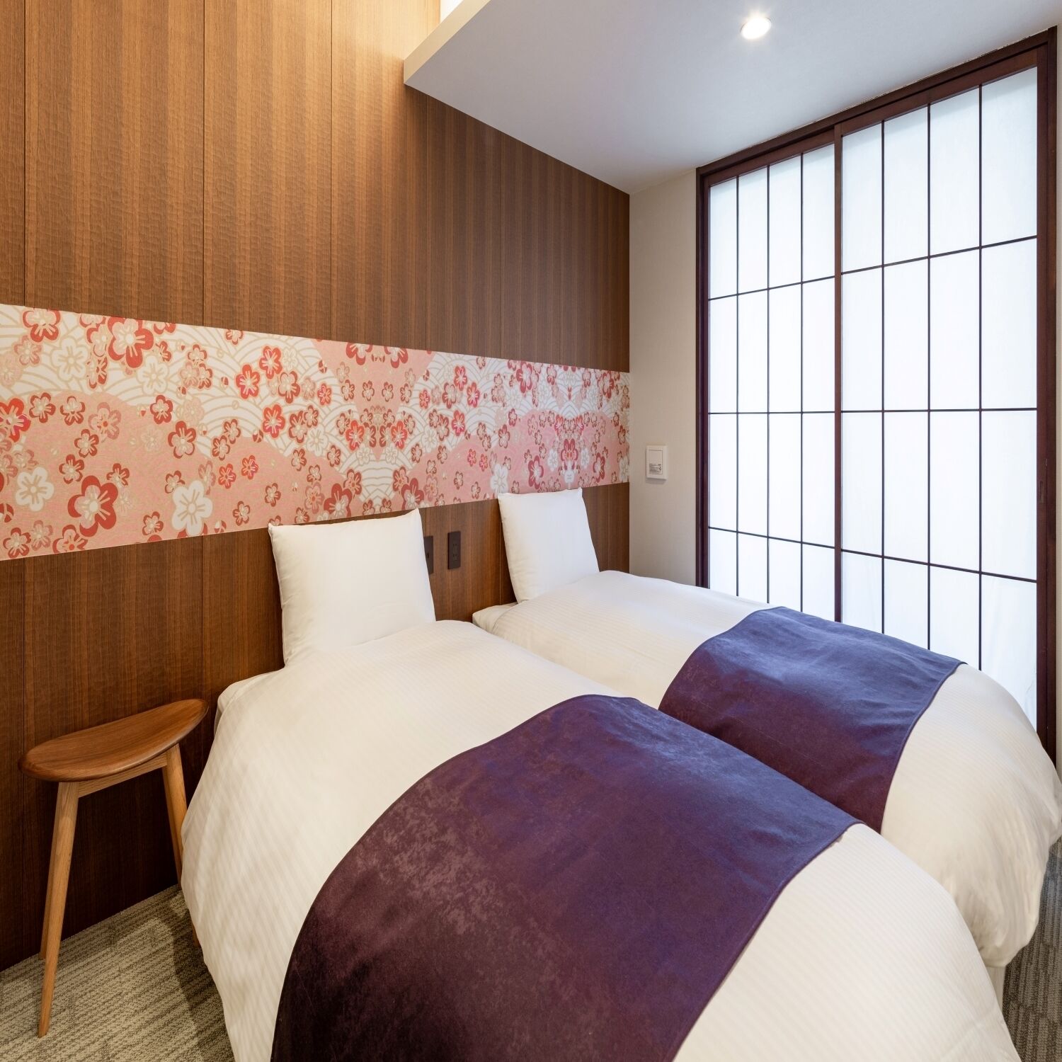 Habitación de hotel con una cama grande, un taburete pequeño, un panel decorativo en la pared y una ventana con diseño de cuadrícula.