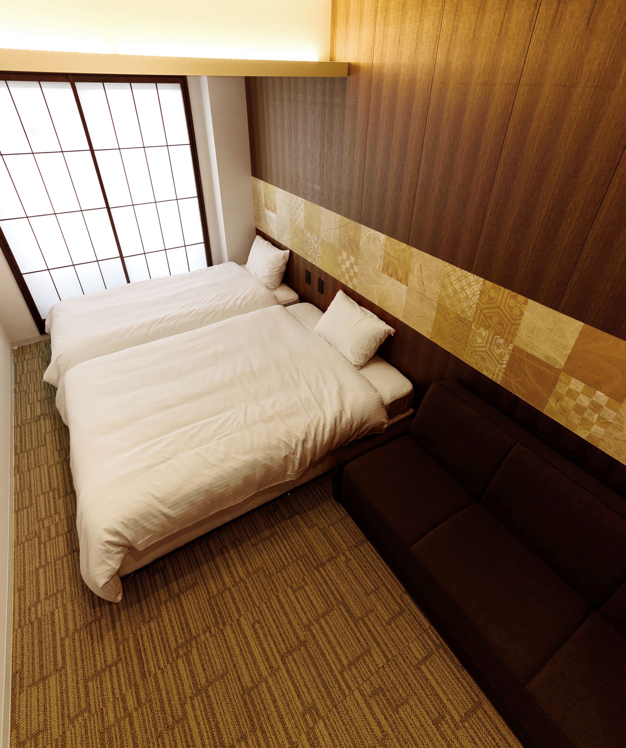 Ein Hotelzimmer mit zwei Betten, einem braunen Sofa, einem gemusterten Teppich und einem hölzernen Kopfteil.