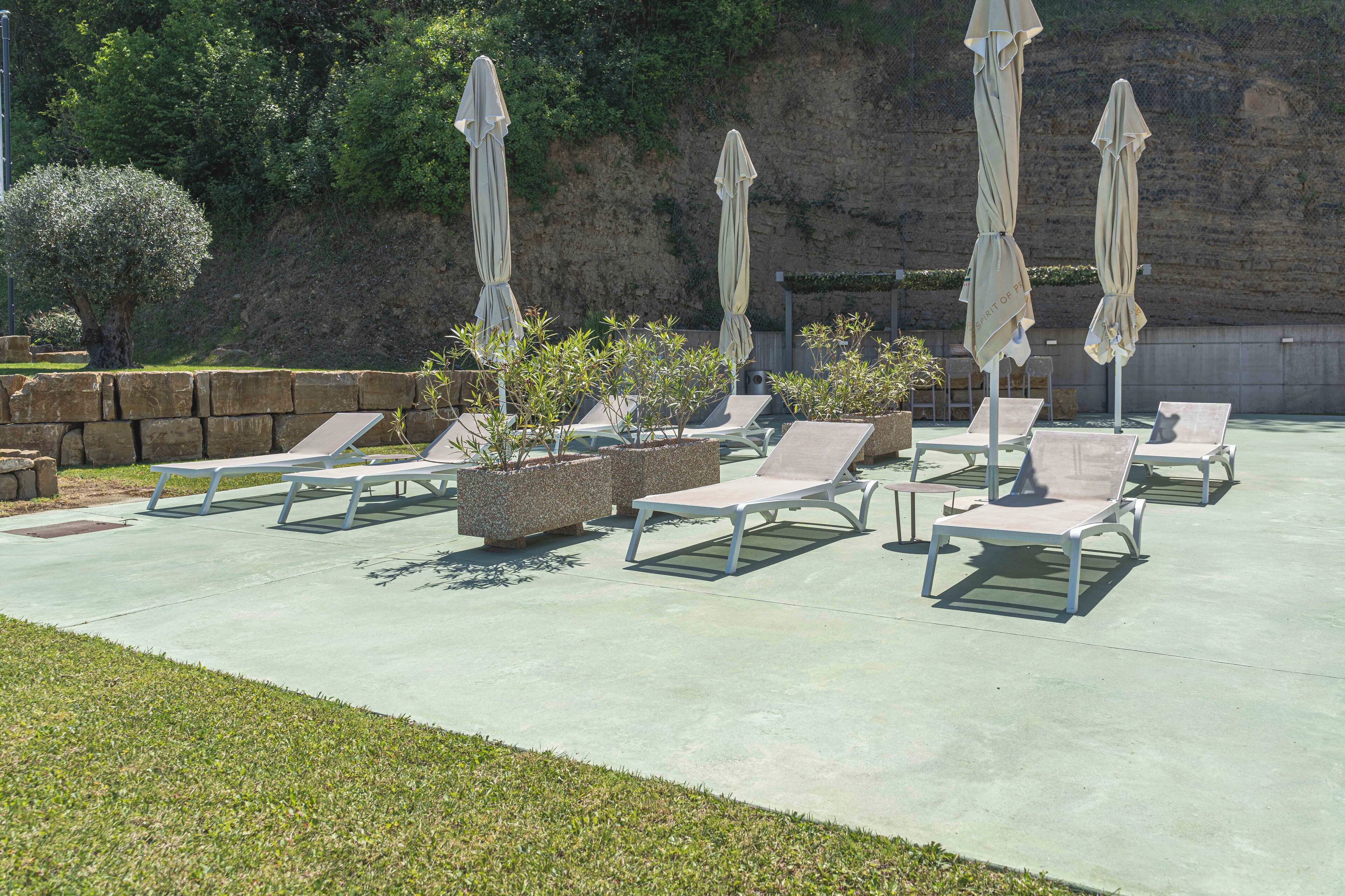 A sunlit outdoor seating area with lounge chairs and umbrellas, surrounded by stone walls and greenery.
