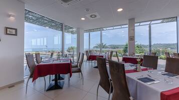 A restaurant with tables set for dining, large windows offering a view of the sea and hills, and a modern interior design.