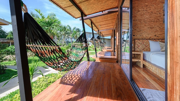 Une terrasse en bois avec un hamac, un banc en bois et un lit doté d’un matelas blanc.