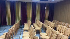 A conference room with rows of beige plastic chairs and tables, a patterned floor, and curtains in gold, maroon, and purple. - Hotel Krishna (Lakhisarai)