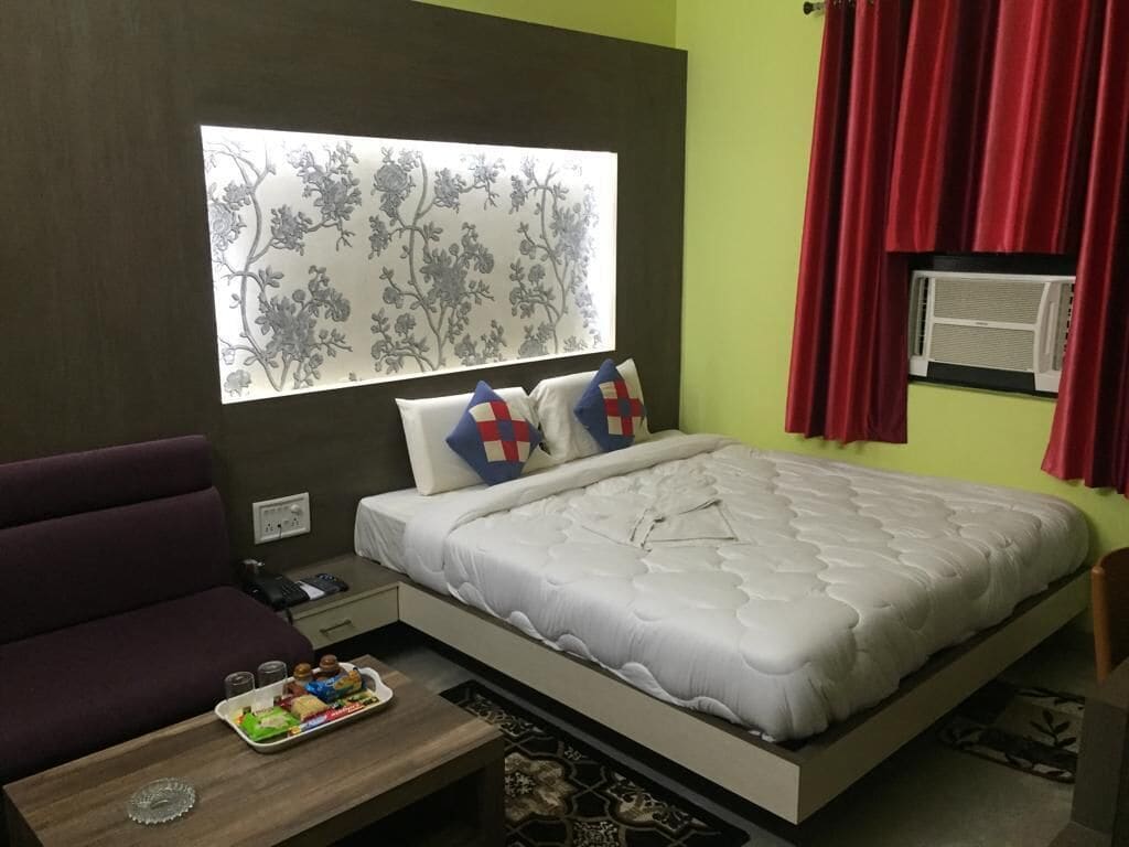 A bedroom with a bed, a sofa, a small table with snacks, a window with a floral design, and an air conditioning unit.