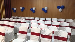 Rows of chairs with white covers and red sashes, arranged in a hall with wooden panel walls and a ceiling with recessed lighting. - Hotel Krishna (Lakhisarai)
