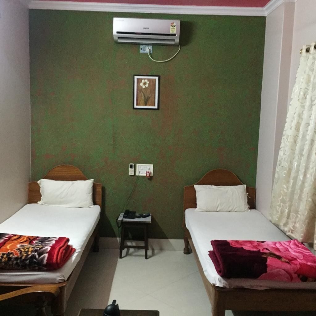 A small room with two single beds, a small table, an air conditioner, and a power outlet on the wall.