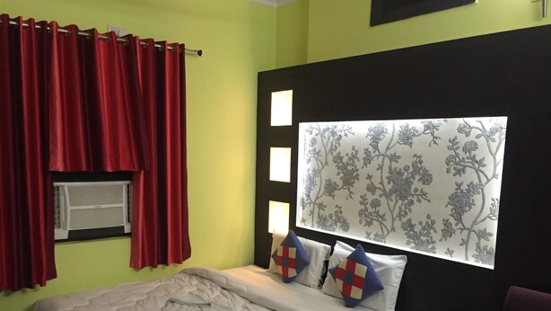 A bedroom with a bed, a nightstand, a lamp, and a window with red curtains. - Hotel Krishna (Lakhisarai)