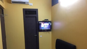 A small room with a dark door, a television mounted on the wall, a black sofa, and a small table with a tray of glasses. - Hotel Krishna (Lakhisarai)