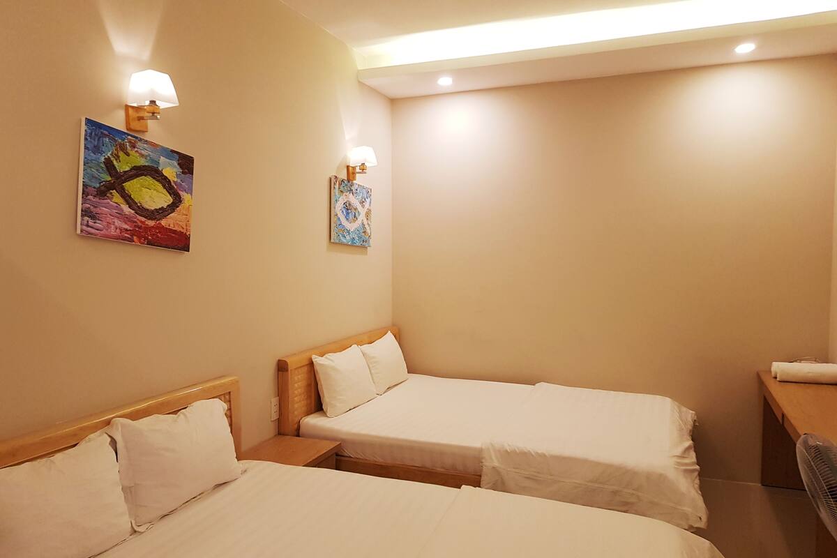 Une chambre d’hôtel avec deux lits simples, une tête de lit en bois, une table de chevet, un bureau et une chaise. Il y a deux lampes fixées au mur et deux œuvres d’art.