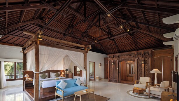 Une pièce spacieuse avec un plafond haut, un grand lit à baldaquin, un coin salon avec un canapé et des fauteuils, et une porte en bois ornée de sculptures complexes.