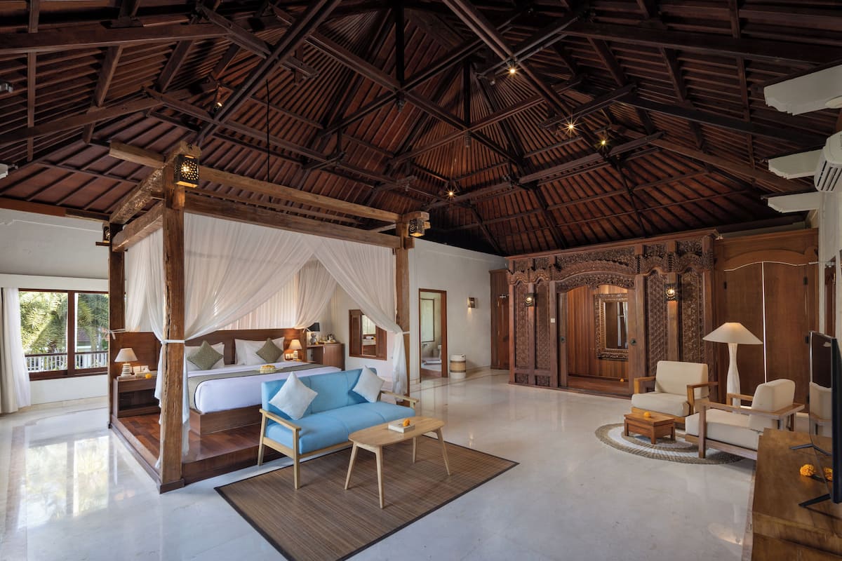 A spacious room with a high ceiling, a large bed with a canopy, a sitting area with a sofa and chairs, and a wooden door with intricate carvings.