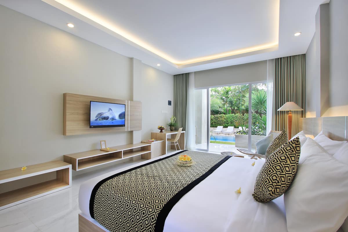 A modern hotel room with a large bed, a TV mounted on the wall, a desk, and a view of the outdoor pool area.