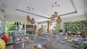 Un vaste espace extérieur aménagé pour les repas, avec de nombreuses tables et chaises, entouré de grandes fenêtres et agrémenté de plantes d’intérieur.