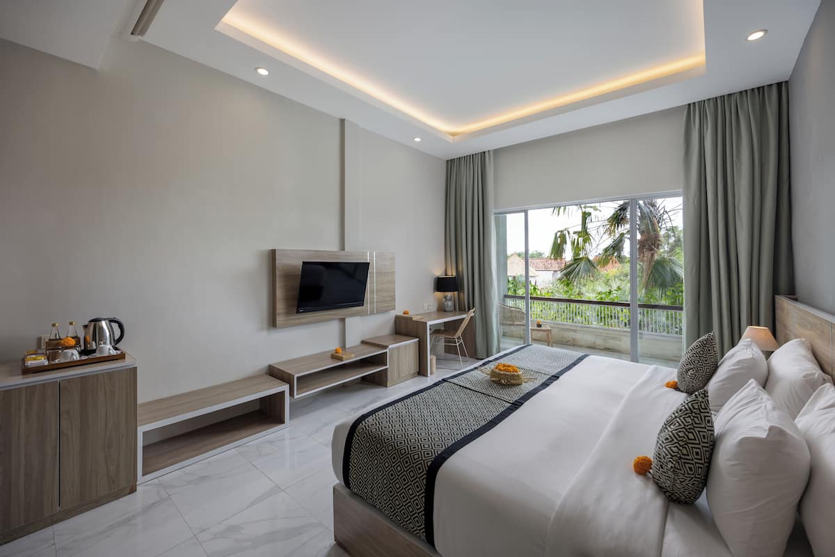 A modern hotel room with a large bed, a TV mounted on the wall, a desk, and a balcony with a view of greenery.