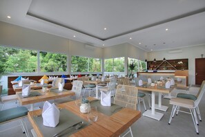 Zona de menjador moderna amb taules de fusta i cadires de vimetlla, grans finestres que ofereixen vistes a la vegetació, i zona de cuina amb una barra i prestatges.