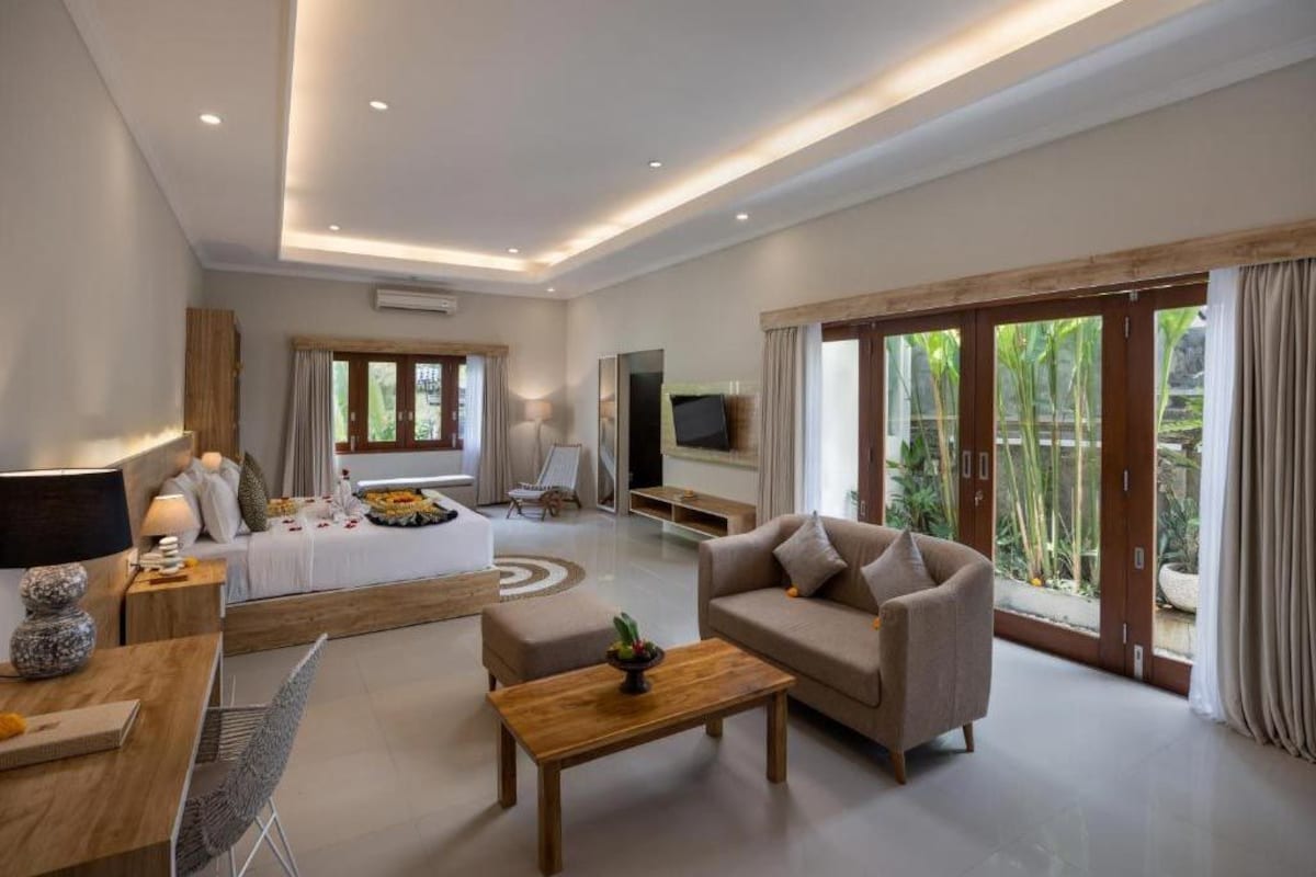 Habitación de hotel moderna con cama, sofá, mesita de café, televisor y grandes puertas correderas de vidrio hacia un jardín.