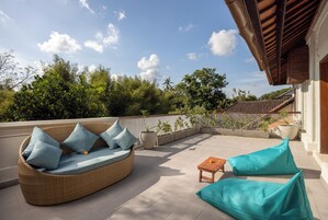 Un divano in vimini con cuscini, un tavolino di legno e due pouf su un patio piastrellato, con vista su alberi e un edificio sullo sfondo.