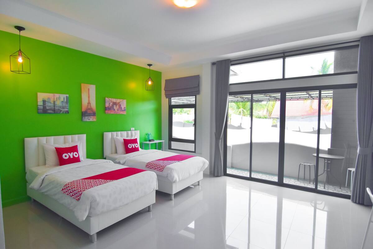 A modern hotel room with two beds, a green accent wall, and large windows offering a view of the outdoors.