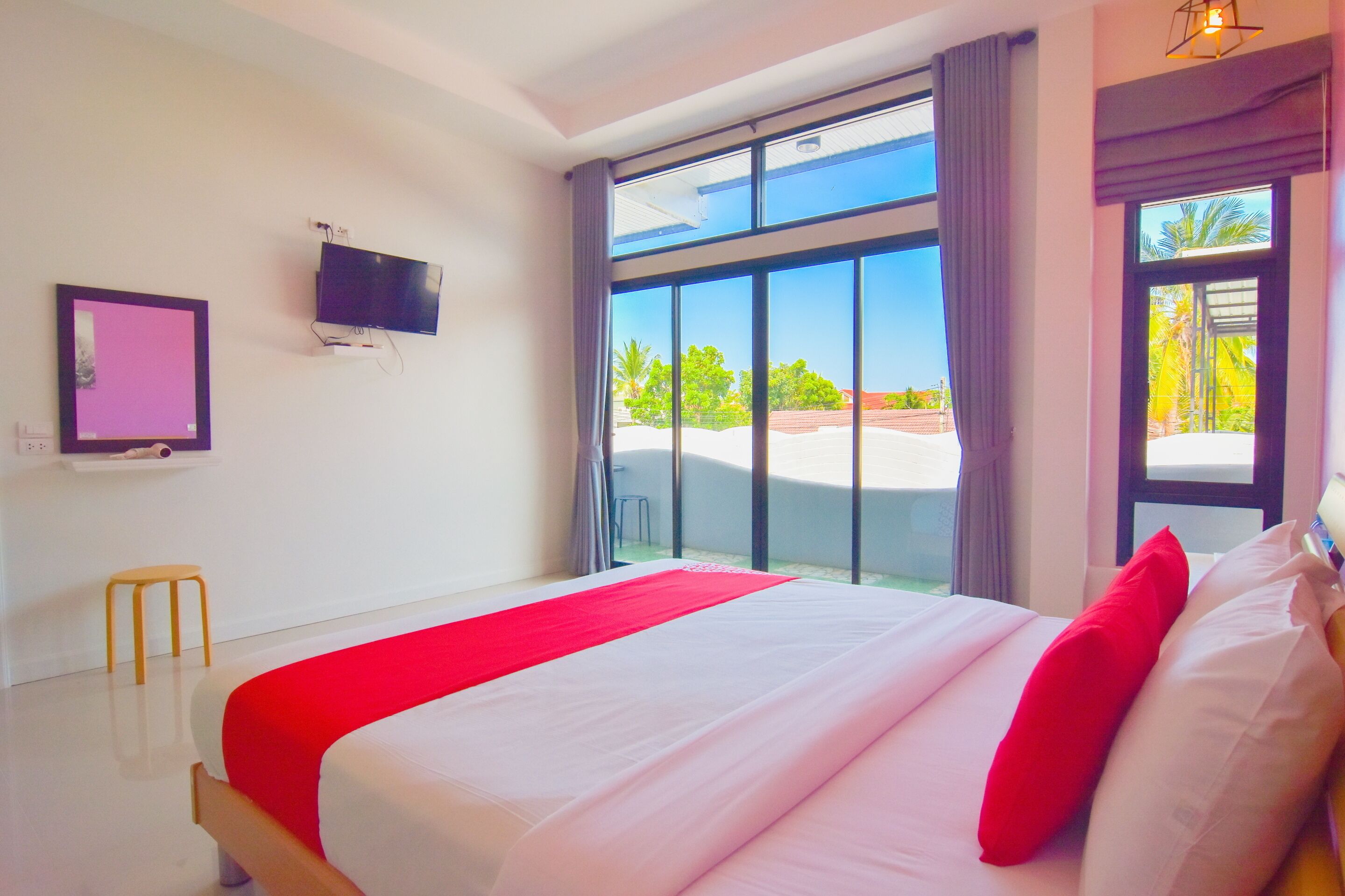 A hotel room with a large bed, a flat-screen TV, a small stool, and a view of the outdoors through large windows.